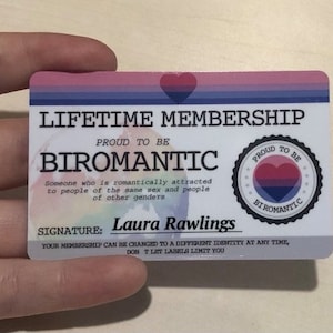 May include: A card with the text "Lifetime Membership" and "Proud to be Biromantic". The card has a pink, blue, and white striped background with a heart symbol in the top left corner. The card also has a smaller circle with the text "Proud to be Biromantic" and a heart symbol in the bottom right corner.