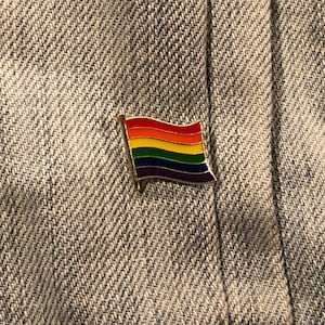 May include: A rainbow flag pin with a gold outline. The pin is attached to a piece of denim fabric.