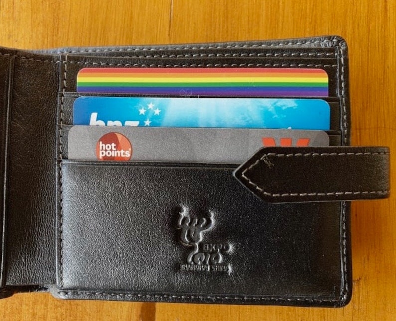 May include: A black leather wallet with a brown leather strap and a logo of a stag. The wallet has several cards inside, including a rainbow card, a blue card with the word "bnz" on it, and a gray card with the words "hot points" on it.