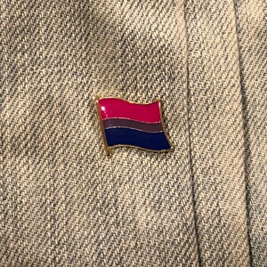 May include: A gold-colored pin with a bisexual pride flag design. The flag has three horizontal stripes: pink, purple, and blue.