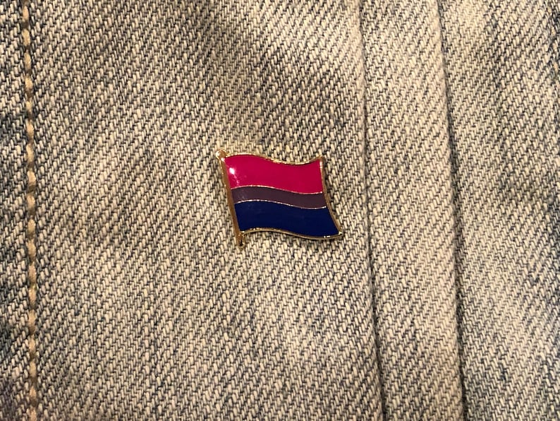 May include: A gold-colored pin with a bisexual pride flag design. The flag has three horizontal stripes: pink, purple, and blue.