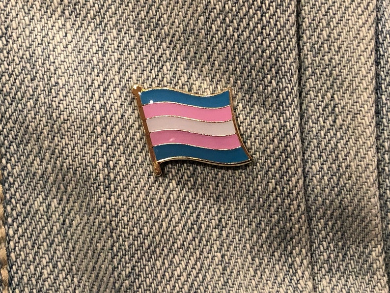 May include: A blue, pink, and white transgender pride flag pin on a denim background.