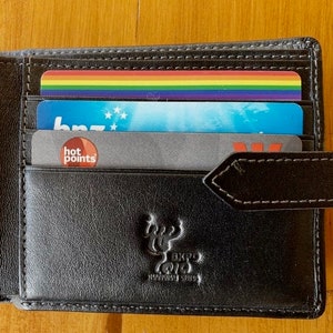 May include: Black leather bi-fold wallet with a snap closure, holding several cards. The cards include a rainbow-colored card, a blue card with white stars, and a gray card with the words "hot points". The wallet has a stitched design and an embossed logo.