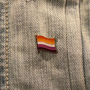 May include: A small, gold-colored pin with a lesbian pride flag design. The flag has five horizontal stripes: orange, white, light pink, white, and dark pink.