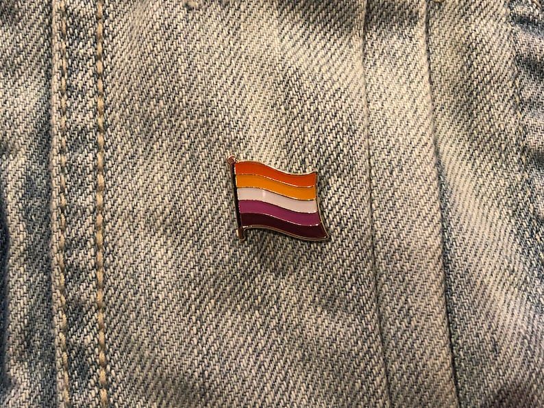 May include: A small, gold-colored pin with a lesbian pride flag design. The flag has five horizontal stripes: orange, white, light pink, white, and dark pink.