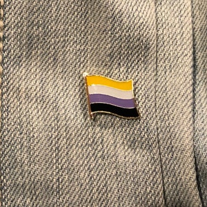 May include: A gold-colored pin with a flag design. The flag has four horizontal stripes: yellow, white, purple, and black. The pin is on a blue denim background.