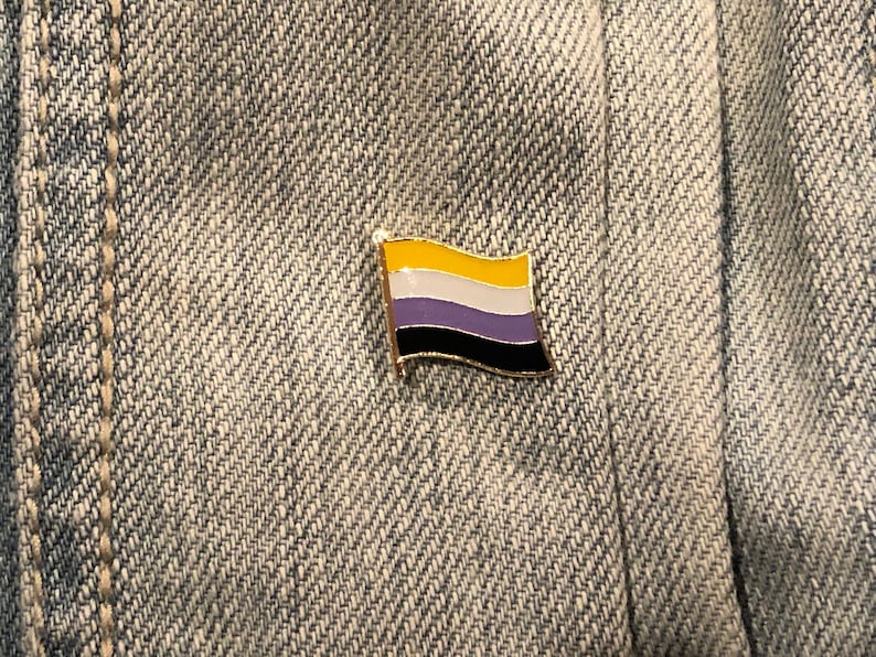 May include: A gold-colored pin with a flag design. The flag has four horizontal stripes: yellow, white, purple, and black. The pin is on a blue denim background.