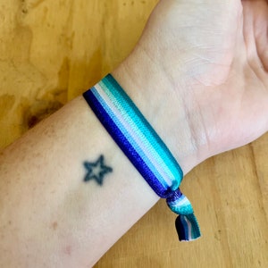 May include: A blue, green, white, and purple striped elastic bracelet with a knot on the end. A small black star tattoo is visible on the wrist.
