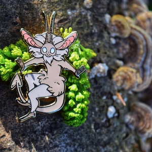 May include: A whimsical enamel pin featuring a light brown creature with a white face, pink ears, and a gold outline. The pin is set against a backdrop of green foliage and a textured tree trunk, creating a nature-inspired aesthetic.