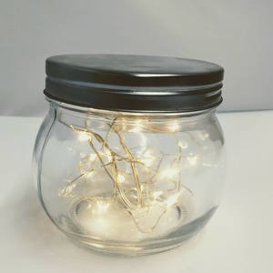 May include: A clear glass jar with a dark grey metal lid. The jar is filled with small, warm white LED string lights. The jar is on a white surface. This decorative jar can be used for home decor.