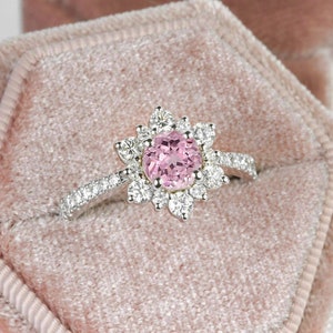 May include: A pink gemstone ring with a halo of smaller diamonds set in a white gold band. The ring is displayed in a pink velvet ring box.