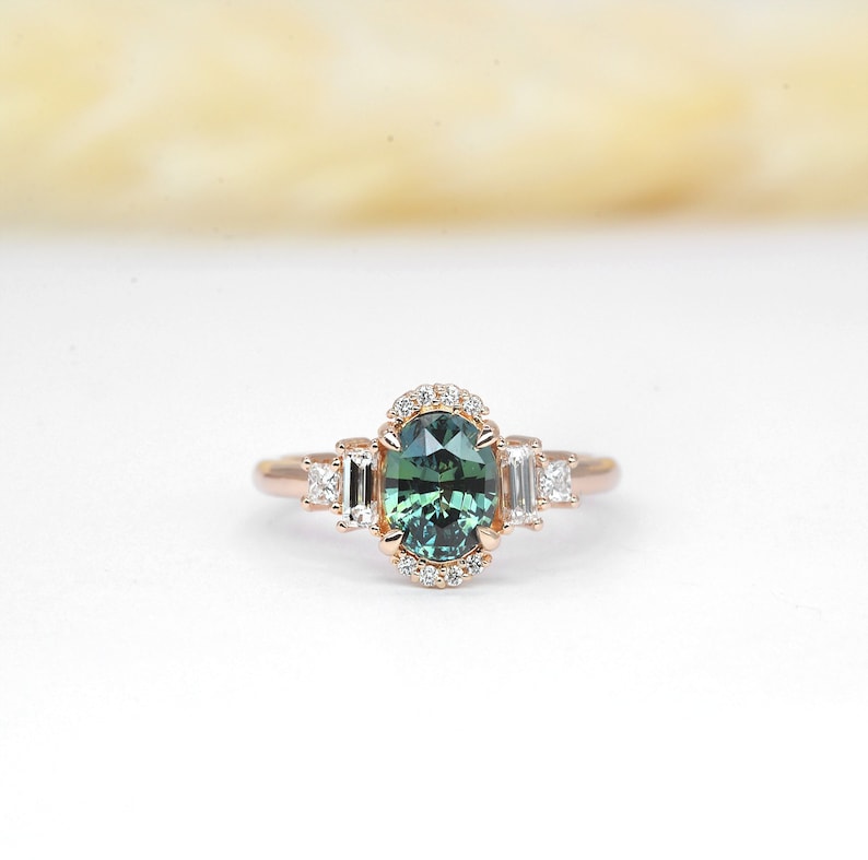 Puede incluir: Un anillo de compromiso de oro rosa con una piedra ovalada verde rodeada por un halo de diamantes m&aacute;s peque&ntilde;os. Tres diamantes de talla baguette est&aacute;n engastados a cada lado de la piedra central.