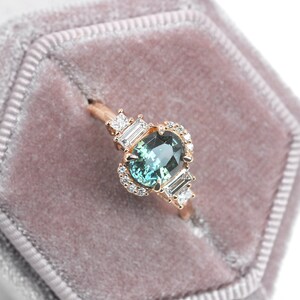 Puede incluir: Un delicado anillo de compromiso con una piedra preciosa verde rodeada por un halo de diamantes m&aacute;s peque&ntilde;os. El anillo est&aacute; engastado en oro rosa y presenta dos diamantes talla baguette a cada lado de la piedra central.