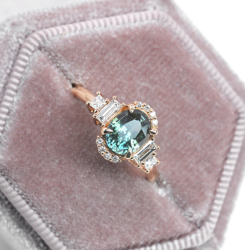 Puede incluir: Un delicado anillo de compromiso con una piedra preciosa verde rodeada por un halo de diamantes m&aacute;s peque&ntilde;os. El anillo est&aacute; engastado en oro rosa y presenta dos diamantes talla baguette a cada lado de la piedra central.