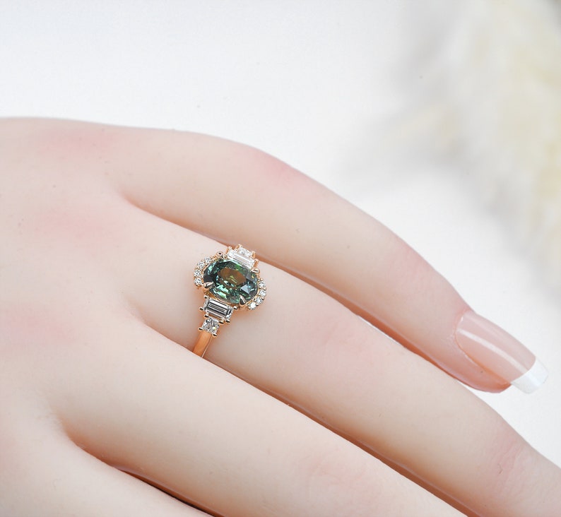 Puede incluir: Un anillo de oro con una piedra ovalada verde rodeada de diamantes m&aacute;s peque&ntilde;os. El anillo est&aacute; puesto en un dedo de maniqu&iacute;.