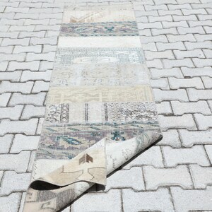 Pode incluir: Um tapete retangular multicolorido com um design de patchwork. O tapete apresenta vários padrões e cores, incluindo bege, azul e creme. O tapete está parcialmente enrolado, revelando a parte inferior. É colocado sobre uma superfície de tijolos cinza claro.