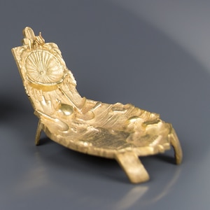 Op de afbeelding: Een gouden metalen sculptuur van een gestileerde stoel met een getextureerd oppervlak. De stoel heeft een ronde rugleuning en drie poten.
