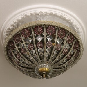 Può includere: Lampada da soffitto ornata con un design decorativo. La luce presenta una combinazione di cristalli trasparenti e rosa, con una base color oro. L'apparecchio è montato su un soffitto bianco con modanature decorative.