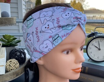 May include: A light blue fabric headband featuring a repeating pattern of cartoon teeth, toothbrushes, and dental floss containers. The headband has a knotted detail at the front and is displayed on a mannequin head with brown hair.