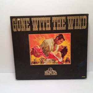 Puede incluir: Una caja de cinta VHS negra con el título en oro "Gone With The Wind" y una imagen de una pareja abrazándose. La caja tiene el logotipo de MGM/UA Home Video.