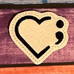 May include: White heart-shaped patch with a black outline and a black semicolon inside. The patch is sewn onto a purple and blue striped fabric.