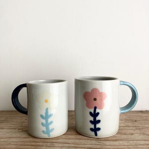 May include: Two white ceramic mugs with floral designs. One mug has a dark blue handle and a white flower with a yellow center and light blue stem. The other mug has a light blue handle and a pink flower with a dark blue stem.