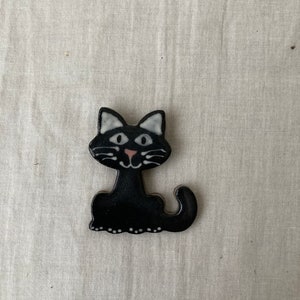 Puede incluir: Figura de gato de cer&aacute;mica negra con rasgos faciales blancos y una nariz rosa. El gato est&aacute; sentado con la cola enroscada alrededor de sus patas.