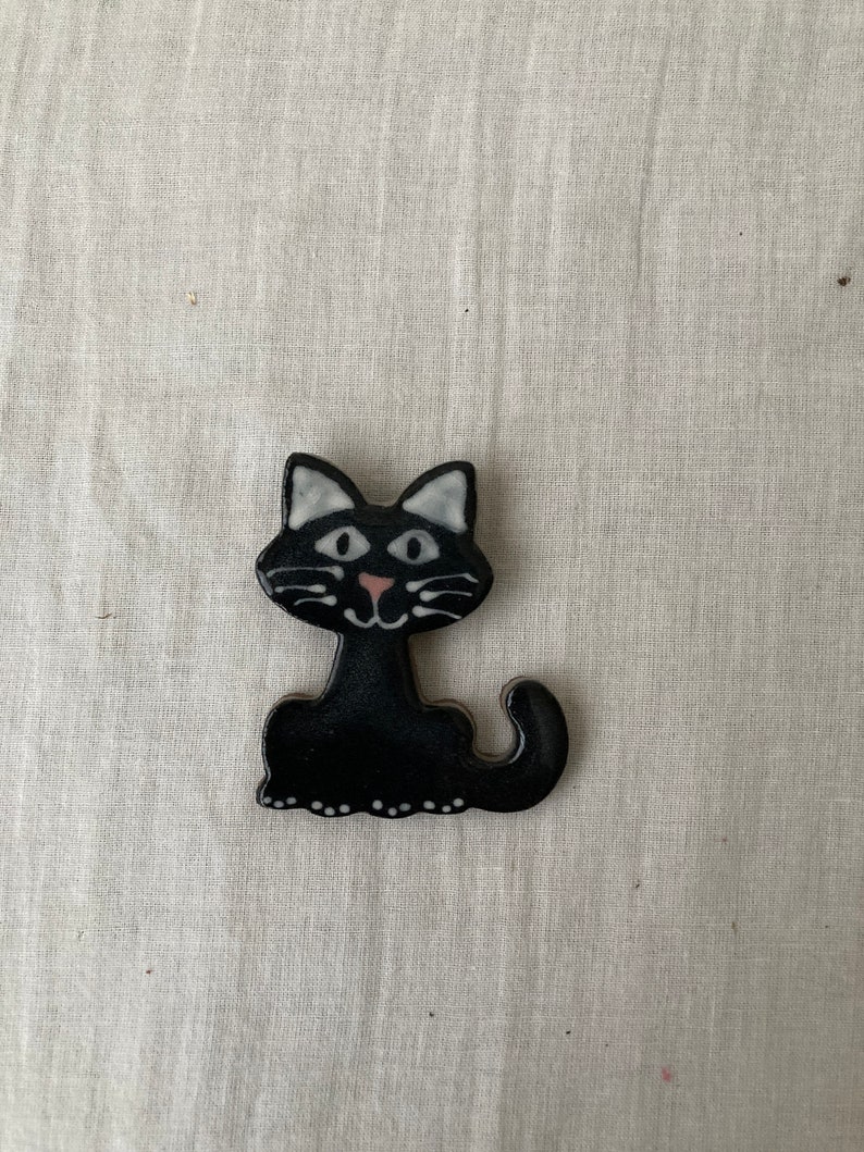 Puede incluir: Figura de gato de cer&aacute;mica negra con rasgos faciales blancos y una nariz rosa. El gato est&aacute; sentado con la cola enroscada alrededor de sus patas.