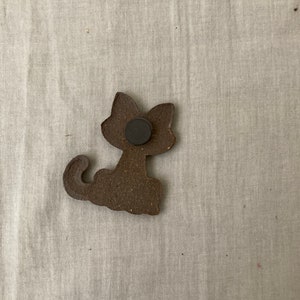 Puede incluir: Una figurita de gato de cer&aacute;mica marr&oacute;n con un im&aacute;n negro en la parte posterior.