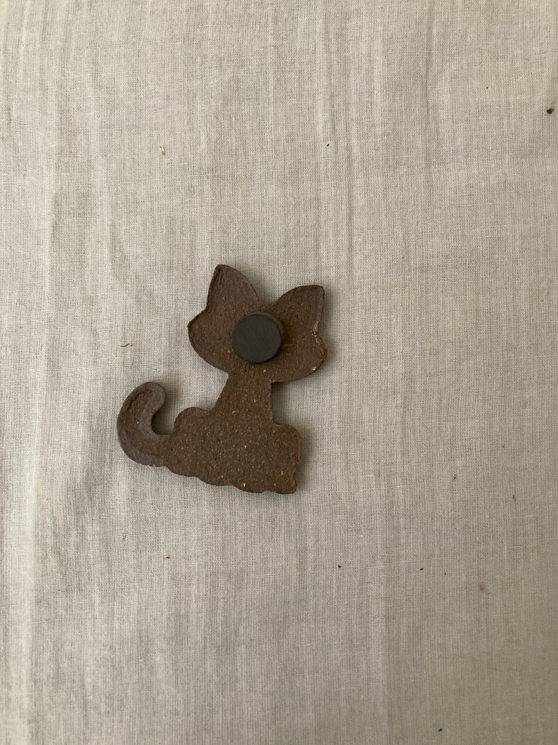 Puede incluir: Una figurita de gato de cer&aacute;mica marr&oacute;n con un im&aacute;n negro en la parte posterior.