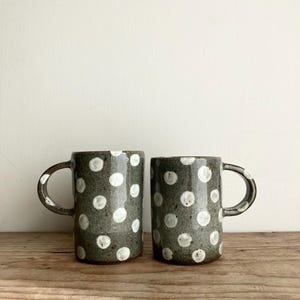 May include: Two ceramic mugs with a dark gray speckled finish and white polka dots. Each mug has a curved handle. The mugs are displayed on a wooden surface against a white background.