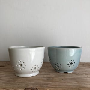 May include: Two ceramic colanders, one white and one light blue, with a decorative pattern of small circles. The colanders are sitting on a wooden surface.