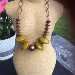 May include: A necklace with a copper chain and olive-green and copper-colored beads. The necklace is displayed on a mannequin. The necklace has a unique design with oval and round beads.