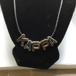 Puede incluir: Un collar con la palabra "KAPPA" escrita con letras de metal doradas y plateadas. Las letras están ensartadas en un fino cordón negro. El collar se exhibe sobre un busto de terciopelo negro.