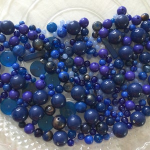 May include: A collection of blue and purple glass beads in various shapes and sizes. The beads are arranged in a random pattern on a white surface.