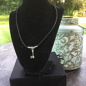 May include: A silver axe pendant necklace on a black display. The pendant hangs from a thin black cord. The axe has a detailed design. The display is on a black surface, with a decorative green lantern in the background.