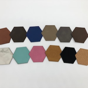 Könnte beinhalten: Eine Sammlung von zwölf sechseckigen Untersetzern in verschiedenen Farben und Materialien. Die Untersetzer umfassen Brauntöne, Blau, Grau, Rosa und Schwarz, mit Materialien wie Holz, Marmor und Kork. Sie sind auf einer weißen Oberfläche angeordnet.