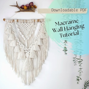 Puede incluir: Un tapiz de pared de macramé color crema con un diseño geométrico y múltiples borlas. La decoración de la pared está suspendida de un pasador de madera. La imagen también incluye el texto "Downloadable PDF" y "Macrame Wall Hanging Tutorial".