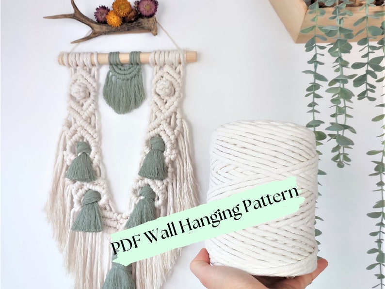 Puede incluir: Un cord&oacute;n de cuerda de algod&oacute;n blanco con una etiqueta verde que dice "PDF Wall Hanging Pattern". La cuerda tiene forma cil&iacute;ndrica y est&aacute; siendo sostenida por una mano. En el fondo, una pared de macram&eacute; con borlas verdes.