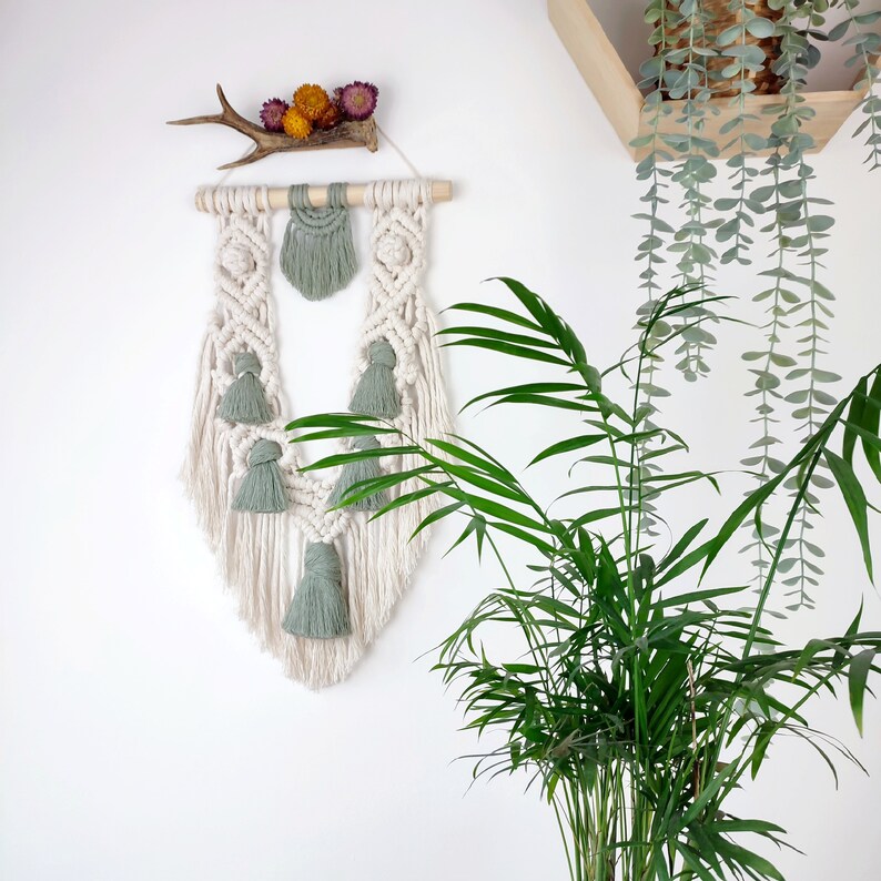 Puede incluir: Un tapiz de macram&eacute; blanco con borlas verdes. El tapiz tiene forma de l&aacute;grima y tiene una varilla de madera en la parte superior. El tapiz est&aacute; montado en una pared blanca con una planta verde en el fondo.