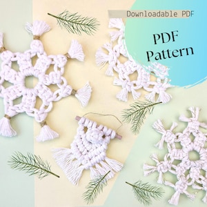 May include: White macrame snowflake decorations with gold detailing and tassels, plus a small macrame wall hanging. The image includes text that reads "Downloadable PDF" and "PDF Pattern". Green sprigs are scattered around the decorations.