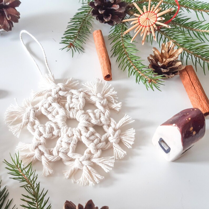 May include: A white crocheted snowflake ornament with a macrame design. The ornament is hanging from a white string and is surrounded by pine branches, cinnamon sticks, and other festive decorations.