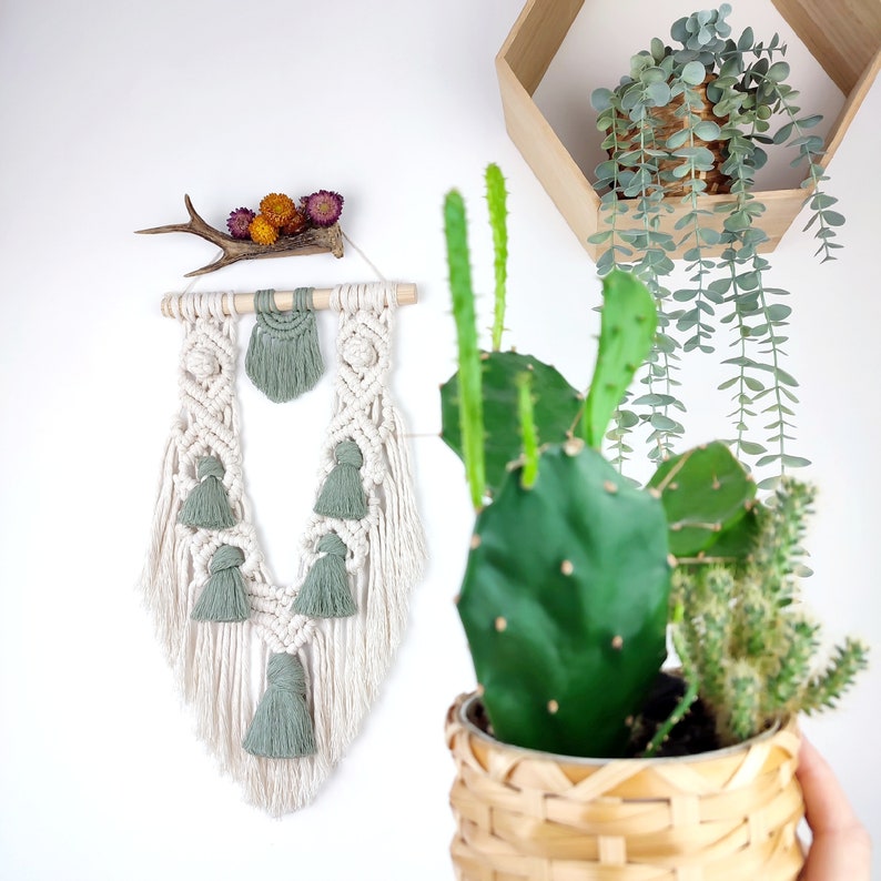 Puede incluir: Un tapiz de macram&eacute; blanco con borlas verdes y una barra de madera. El tapiz tiene forma de V y tiene una cornamenta natural con flores secas en la parte superior. El tapiz se muestra en una pared blanca con un cactus en maceta en primer plano.