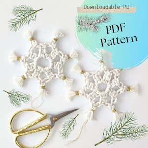 Puede incluir: Dos adornos de macramé en forma de copo de nieve blanco con borlas doradas. Los copos de nieve están sobre un fondo blanco con ramitas de pino verdes y tijeras doradas.