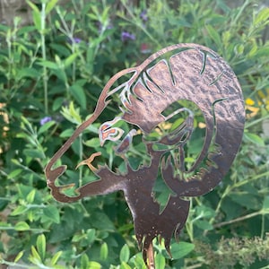 May include: A metal garden stake featuring a ram's head silhouette. The copper-colored stake has intricate cut-out details and is set against a backdrop of green foliage and purple flowers. The ram's head is facing left.