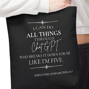 May include: Black tote bag with a burgundy strap. The bag features white text that reads "I CAN DO ALL THINGS THROUGH ChatGPT WHO BREAKS IT DOWN FOR ME LIKE I'M FIVE. EXECUTIVE DYSFUNCTION 24/7."