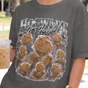 May include: A charcoal gray short-sleeve t-shirt featuring a graphic print of multiple Hoggle characters from the movie Labyrinth. The text at the top reads HOGWARTS It's Hoggle! in a metallic silver font, surrounded by a cloudy, lightning-like effect.