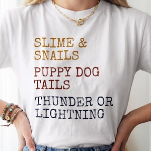 May include: A white short-sleeved t-shirt featuring the text SLIME & SNAILS in mustard yellow, PUPPY DOG TAILS in rust brown, and THUNDER OR LIGHTNING in dark navy blue. The lettering is printed in a distressed, block-style font on the front.