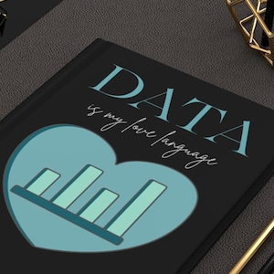 May include: A black journal with the text "DATA is my love language" in teal. A teal heart graphic with a bar graph design is centered on the cover. Gold and black pencils and gold binder clips are also visible.