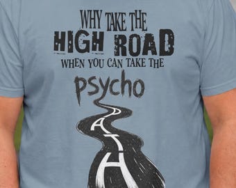 May include: A dusty blue short-sleeve t-shirt featuring black text that reads Why take the high road when you can take the psycho path. The word path is stylized as a winding road graphic with white lane markings running down the center of the road.
